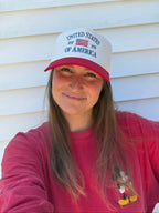 A woman wearing a red and white 'United States of America Est. 1776 ' trucker hat posing in front of a white siding wall.